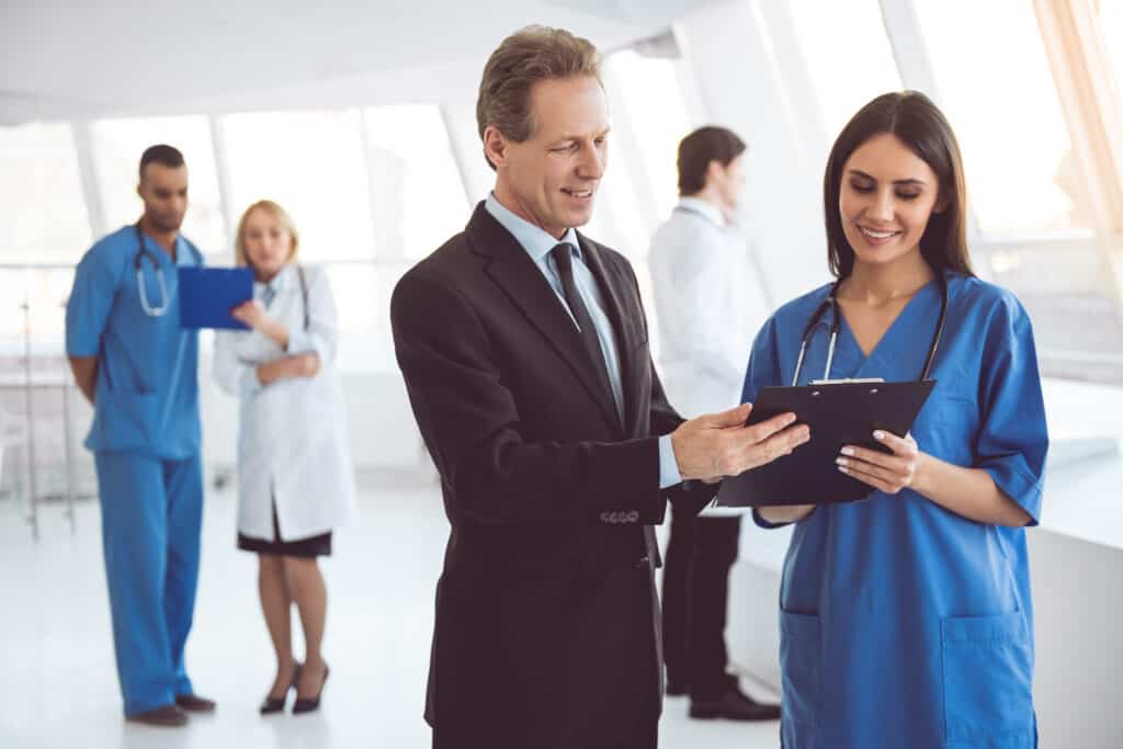 an in a suit speaks with a woman in scrubs while reviewing a clipboard together in a medical setting.