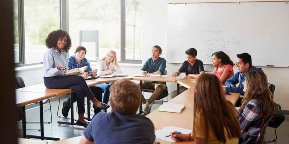 Students are sitting at desks arranged in a circle. Their teacher is sitting on one of the desks and giving a lecture.