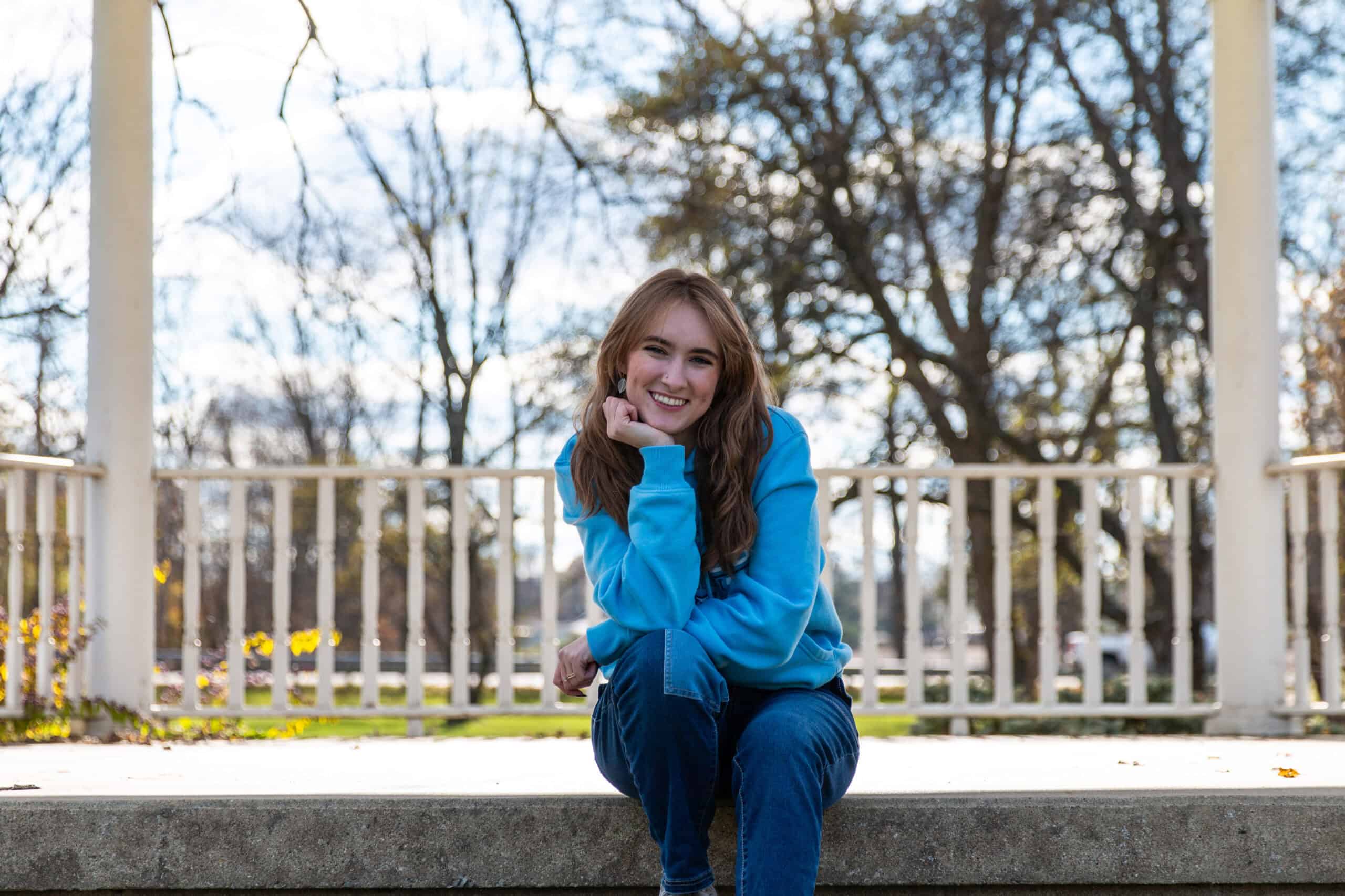 A young woman with long brown hair wearing a light blue hoodie and jeans sits on a stone ledge, smiling brightly with her head resting on her hand. She is outdoors in a park-like setting with bare trees and a white railing behind her, under a partly cloudy sky.