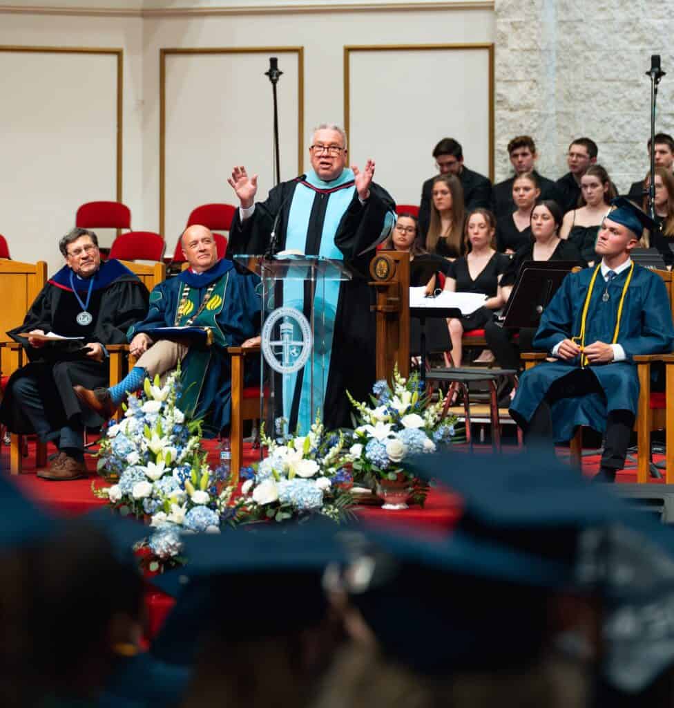 man in black robe with blue sash speaks to graduates