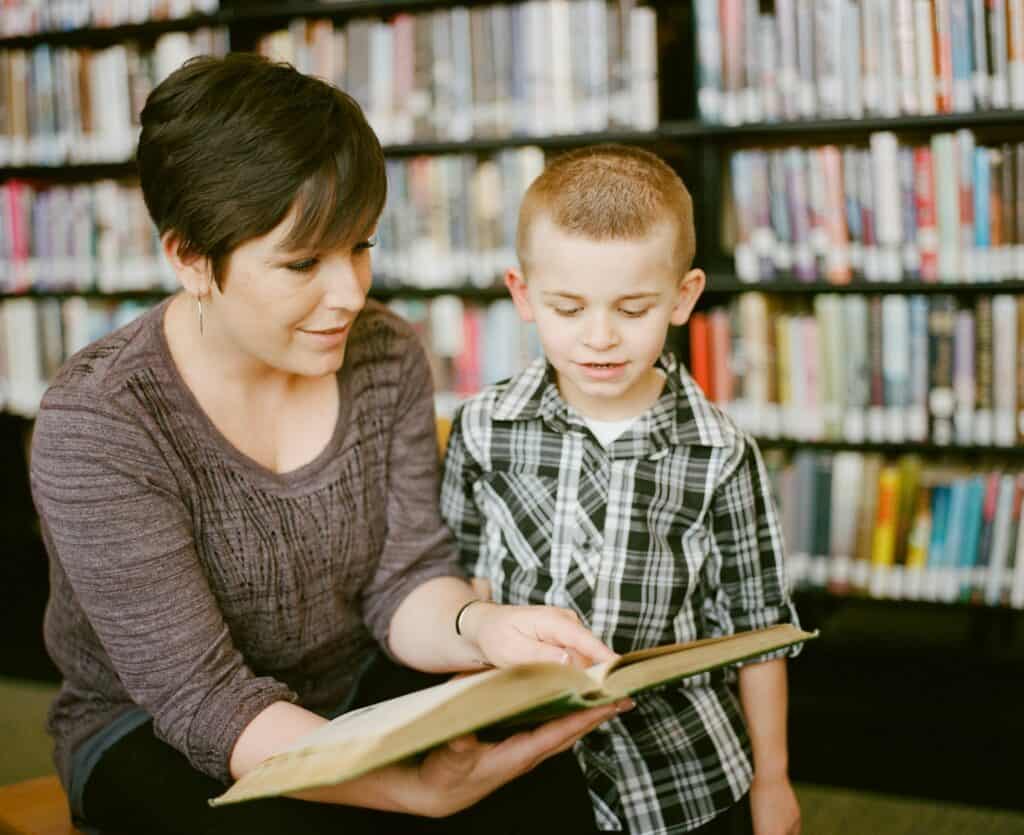 Adult helping young child read a book in a library, representing the impact of earning an online teaching degree in Cleveland.