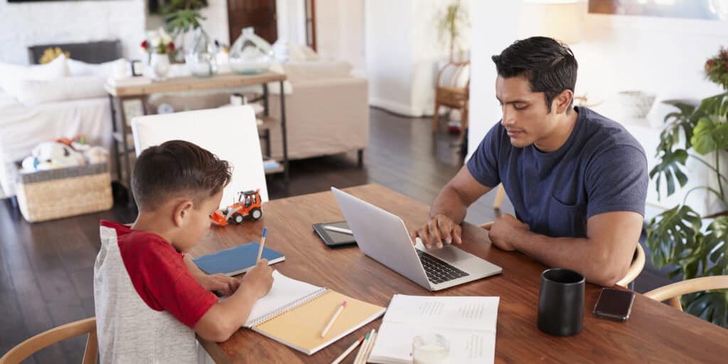 An adult student working on a laptop at the kitchen table alongside his child doing homework—highlighting how MVNU Online supports busy parents balancing school and family.