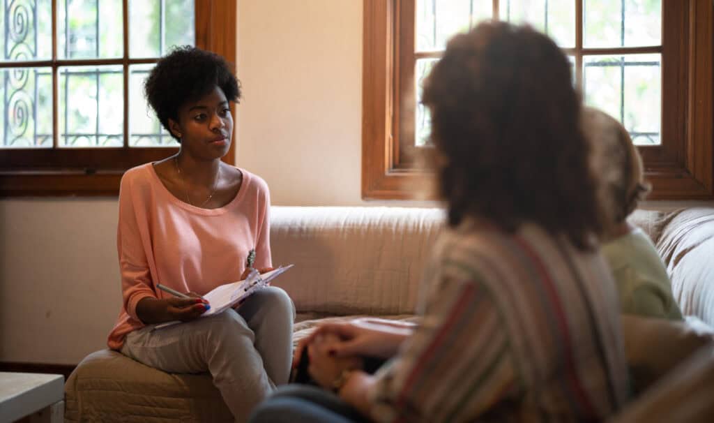 A young woman holding a clipboard listens attentively to two people during a counseling session, representing compassionate, faith-informed social work.